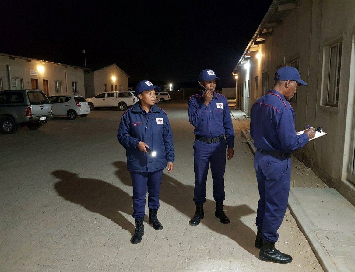 Maya night-rounds team patrolling a lit complex with torch, radio and clipboard